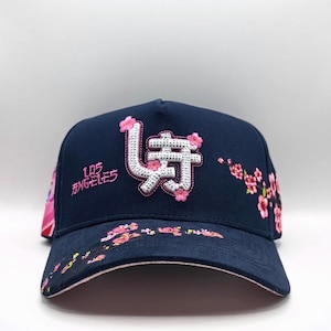 May include: Navy blue baseball cap with embroidered Japanese characters and pink cherry blossoms. The cap features the text "Los Angeles" in pink. The brim and side panels are decorated with floral designs.