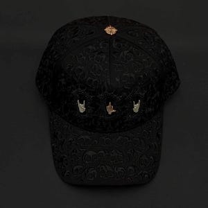 Pode incluir: Um boné de beisebol preto com um padrão de caveiras em relevo por toda a superfície. O painel frontal é adornado com três berloques metálicos de gestos com as mãos em tons de prata e ouro. Um emblema metálico circular está no topo da coroa.