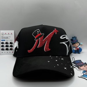 May include: Black baseball cap featuring a red and rhinestone-studded letter "M" with a top hat design. The cap has a curved brim with white star accents and a small illustration of a magician.