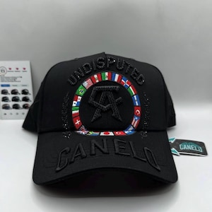 Puede incluir: Gorra de béisbol negra con la palabra "UNDISPUTED" arqueada en la parte delantera, sobre un diseño circular con banderas y las letras "CA". La palabra "CANELO" está bordada en la visera. Una pequeña etiqueta con la palabra "CANELO" está adjunta.