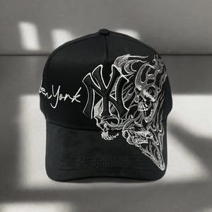 Op de afbeelding: Zwarte baseball cap met het New York Yankees logo en "New York" in witte letters. De cap heeft een zilverkleurig geborduurd schedel- en vlammenontwerp aan de zijkant.