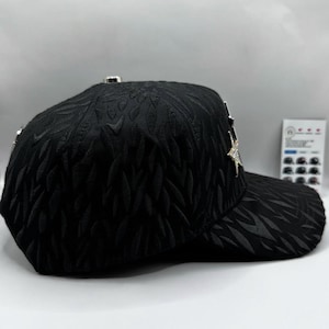 Puede incluir: Gorra de b&eacute;isbol negra con un patr&oacute;n de hojas texturizadas. La gorra presenta un adorno plateado en forma de estrella y detalles plateados. La visera es curva y la gorra tiene un dise&ntilde;o estructurado.