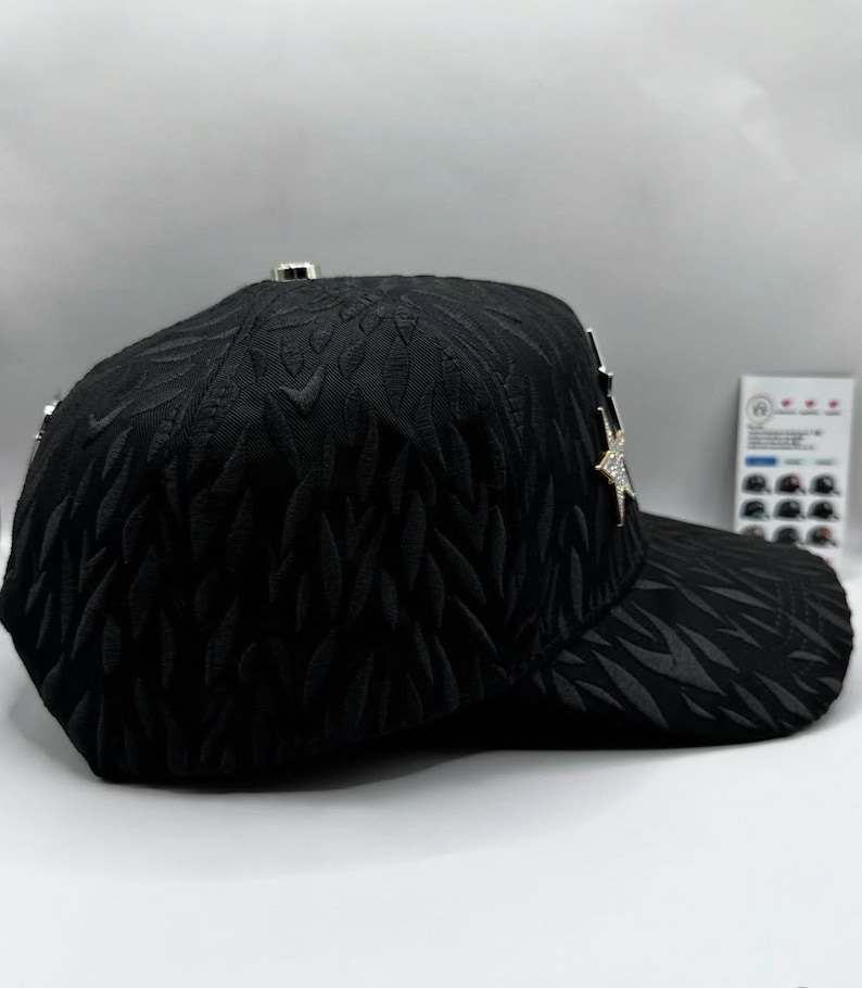 Puede incluir: Gorra de b&eacute;isbol negra con un patr&oacute;n de hojas texturizadas. La gorra presenta un adorno plateado en forma de estrella y detalles plateados. La visera es curva y la gorra tiene un dise&ntilde;o estructurado.