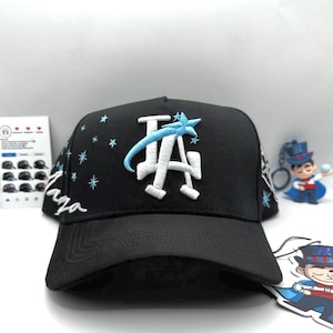 Może przedstawiać: Czarna czapka z daszkiem z białym haftowanym logo "LA" i niebieskim wzorem gwiazd. Czapka ma zakrzywiony daszek i strukturalną koronę. Słowo "Laga" jest wyhaftowane z boku na biało.