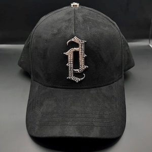 Puede incluir: Gorra de b&eacute;isbol negra con textura similar a la gamuza. La gorra presenta una letra "A" plateada y ornamentada, adornada con peque&ntilde;as piedras brillantes. Un peque&ntilde;o acento plateado est&aacute; en la parte superior de la gorra.