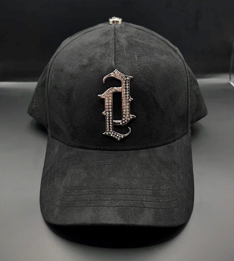 Puede incluir: Gorra de b&eacute;isbol negra con textura similar a la gamuza. La gorra presenta una letra "A" plateada y ornamentada, adornada con peque&ntilde;as piedras brillantes. Un peque&ntilde;o acento plateado est&aacute; en la parte superior de la gorra.