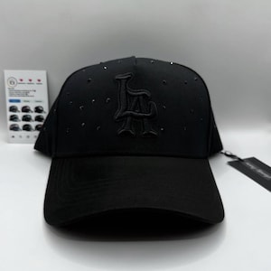 May include: Black baseball cap with the letters "LA" embroidered on the front. The cap has a curved brim and small embellishments. A tag is attached to the side.