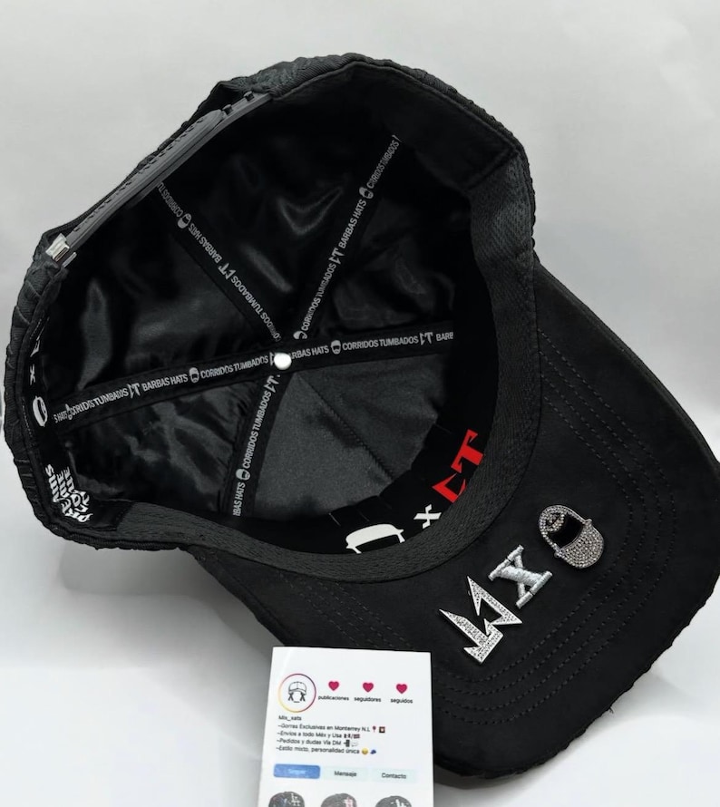 Puede incluir: Gorra de b&eacute;isbol negra con el texto "MX" en pedrer&iacute;a plateada. La gorra tiene una visera curva y un forro de sat&eacute;n negro con texto blanco. Tambi&eacute;n tiene el texto "BARBAS HATS CORRIDOS TUMBADOS IT BARE".