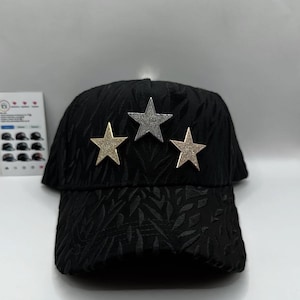 Puede incluir: Gorra de b&eacute;isbol negra con un patr&oacute;n texturizado y tres adornos de estrellas. Las estrellas son doradas y plateadas, a&ntilde;adiendo un toque de brillo al sombrero. La gorra tiene una visera curva.