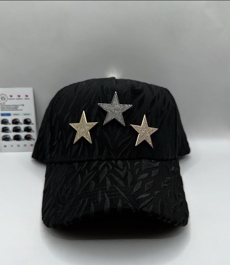 Puede incluir: Gorra de b&eacute;isbol negra con un patr&oacute;n texturizado y tres adornos de estrellas. Las estrellas son doradas y plateadas, a&ntilde;adiendo un toque de brillo al sombrero. La gorra tiene una visera curva.
