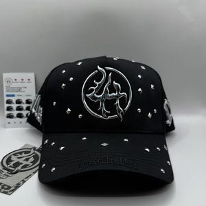 May include: Black baseball cap with silver embellishments. The cap features a silver emblem on the front and is studded with silver accents. The brim has the text "lastrallyx". A tag with a matching emblem is attached.