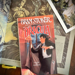 Może przedstawiać: Miękka okładka książki Brama Stokera „Dracula”, klasycznej powieści grozy gotyckiej. Okładka przedstawia dramatyczną ilustrację mężczyzny w pelerynie, stojącego w pokoju z oknem. Tekst „Wierz w niego i módl się, żeby cię nigdy nie znalazł...” jest wydrukowany nad tytułem.
