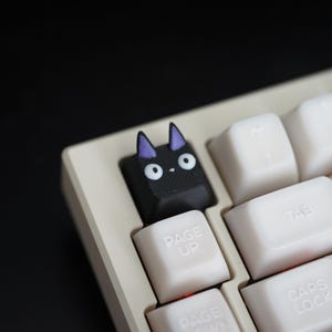 Pode incluir: Uma tecla de teclado personalizada preta com formato de gato e orelhas roxas. A tecla está instalada em um teclado creme com teclas brancas. A tecla do gato tem olhos brancos e um nariz pequeno.