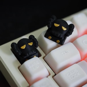 May include: Two black keycaps with yellow eyes and accents are installed on a white keyboard. The keycaps have a unique design, and the keyboard has a white frame and white keys. The keyboard is lit with a red glow.