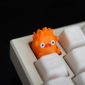 May include: An orange, cartoon-like character keycap sits on a white keyboard. The keycap has a spiky head, round eyes, and a small mouth. The keyboard keys are white with black lettering.
