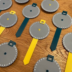 Puede incluir: Un conjunto de nueve diales circulares grises con números impresos alrededor del borde. Cada dial tiene una pequeña pestaña amarilla en la parte superior y un puntero negro en el centro. Los diales están unidos a palos de diferentes colores, incluyendo amarillo, azul verdoso y negro.