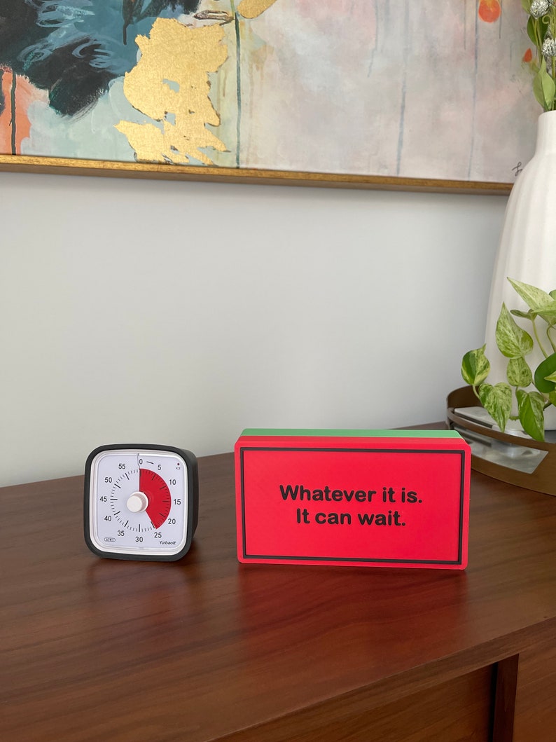 May include: A black and white kitchen timer with a red dial set to 5 minutes. A red and green block with the text "Whatever it is. It can wait." is on a wooden surface.