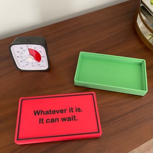 May include: A red rectangular sign with black text that reads "Whatever it is. It can wait." The sign is on a wooden surface next to a green rectangular tray and a black kitchen timer.