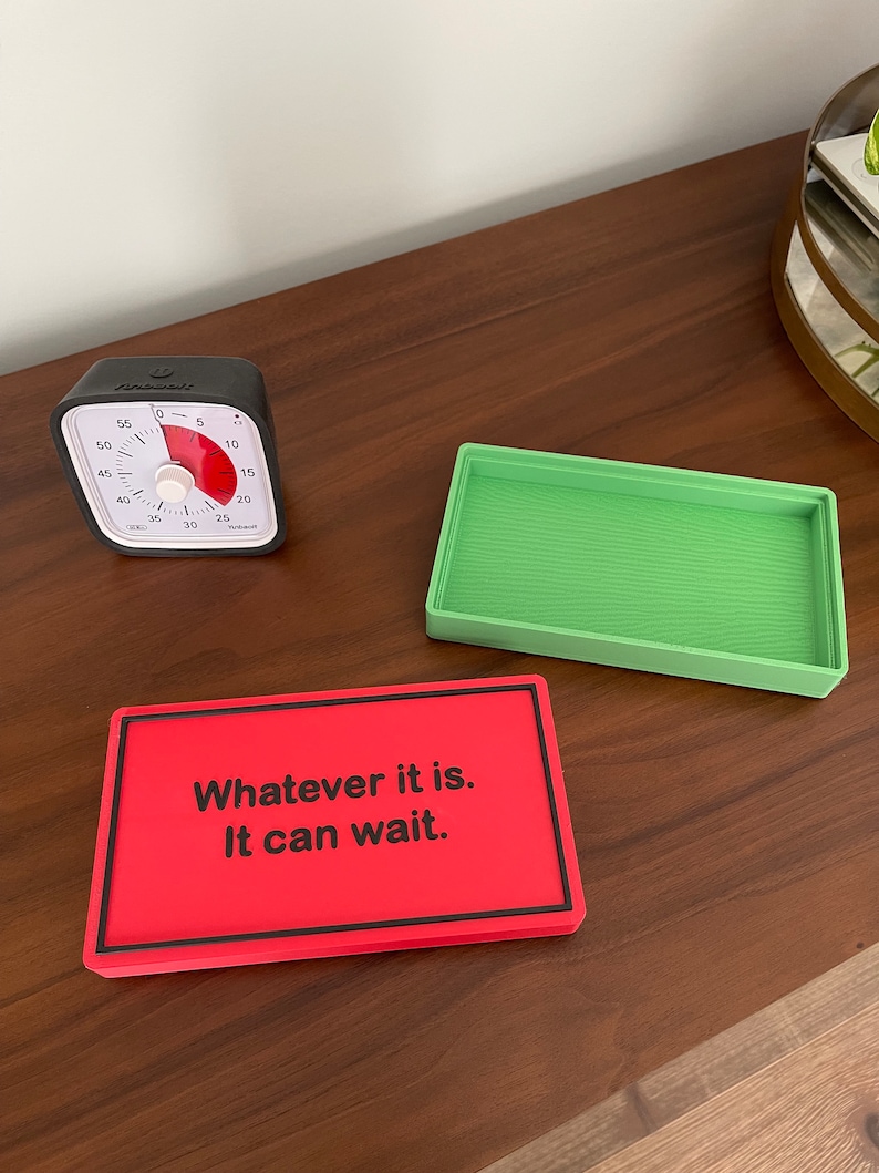 May include: A red rectangular sign with black text that reads "Whatever it is. It can wait." The sign is on a wooden surface next to a green rectangular tray and a black kitchen timer.