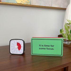 May include: A black and white kitchen timer with a red dial set to 25 minutes. A green and red rectangular sign reads "It is time for some focus."