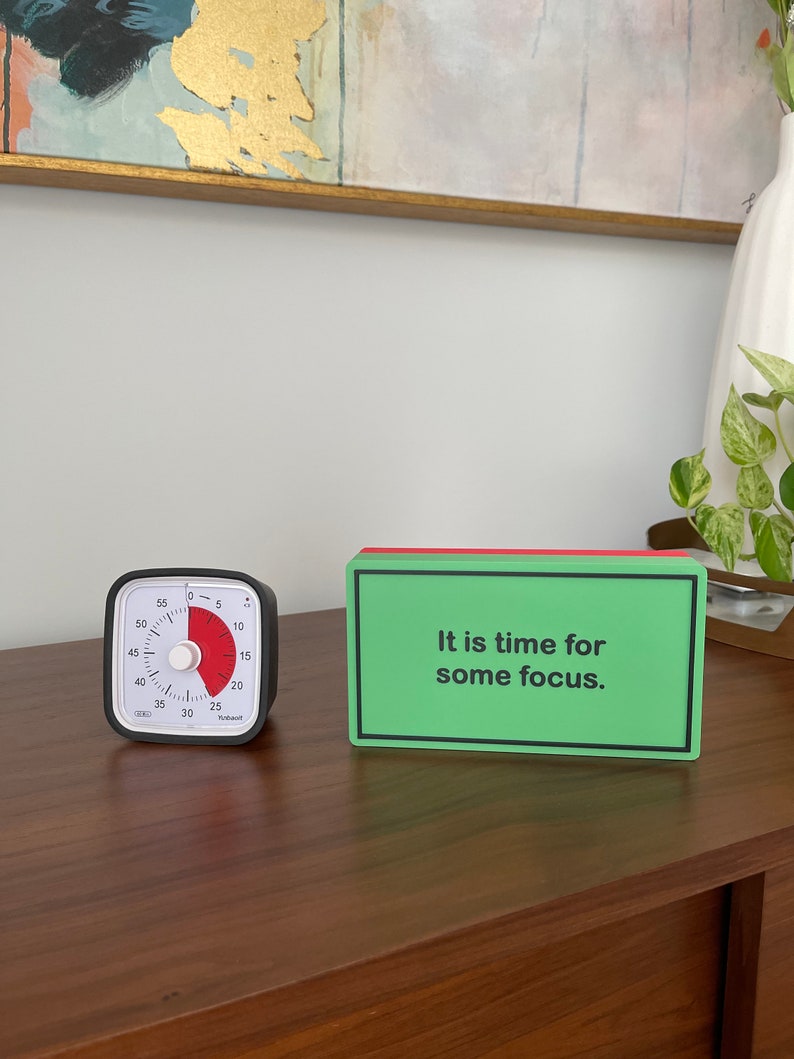 May include: A black and white kitchen timer with a red dial set to 25 minutes. A green and red rectangular sign reads "It is time for some focus."