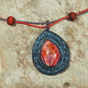 May include: A teal blue pendant necklace with a red flower design inside a clear resin oval. The pendant is attached to a red and gray cord.