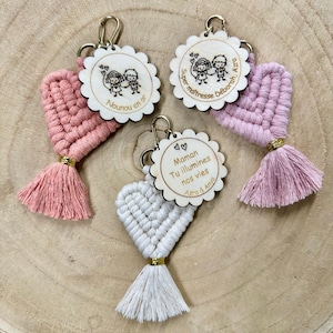 Op de afbeelding: Drie macramé hart-sleutelhangers met goudkleurige metalen sluitingen en kwastjes. Elk exemplaar heeft een ronde houten schijf met geschulpte randen, gegraveerd met illustraties van kinderen en Franse tekst zoals Nounou en or, Super maîtresse Déborah en Maman Tu illumines nos vies.