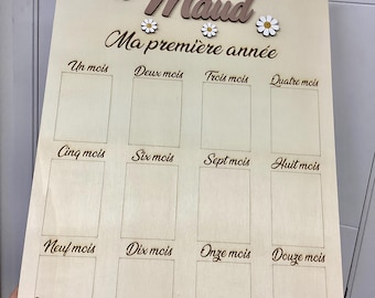 Puede incluir: Un tablero de hitos de madera clara con el nombre Maud y el texto Ma première année. Incluye doce espacios rectangulares para fotos etiquetados desde Un mois hasta Douze mois, decorados con pequeños recortes de margaritas blancas y amarillas.