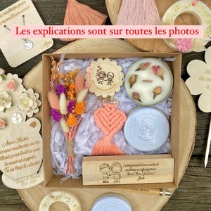 Peut inclure: Un coffret cadeau avec divers articles, dont un porte-clés cœur en macramé pêche, un petit bouquet de fleurs séchées et deux contenants ronds. Le coffret est sur une surface en bois.