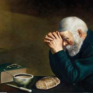May include: An elderly man with white hair and a beard is praying with his hands clasped together. He is wearing a green sweater and is sitting at a table with a book, a bowl, a spoon, and a loaf of bread.