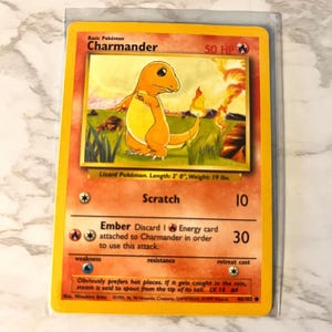May include: A Charmander Pokémon trading card in a protective sleeve. The card features an orange lizard-like creature with a flame on its tail. The card has the text "Charmander" and "50 HP". The card is 2 feet long and weighs 19 pounds.