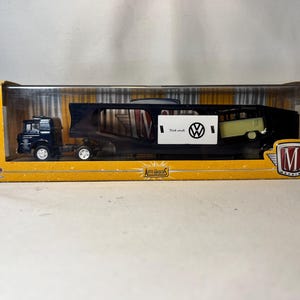 May include: A black semi-truck and trailer carrying a light green and white Volkswagen van. The trailer has a black frame. A white sign with the Volkswagen logo and the words "Think small" is attached to the trailer. The toy is in a yellow box.