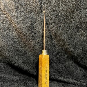 May include: A vintage U.S. Hickory brand rug hook with a wooden handle and a metal hook. The handle is a light brown color and has the brand name printed on it. The hook is made of metal and is curved at the end.