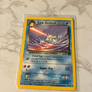 May include: A Dark Golduck Pokémon card in a protective sleeve. The card features a blue and yellow design with an illustration of Dark Golduck using a laser beam. The card's text includes the HP, attack, and other game details.