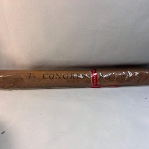 May include: A rolled parchment document encased in clear plastic packaging. The document features the printed text IN CONGRESS, JULY 4, 1776. A small red paper band with white text wraps around the center of the rolled document.