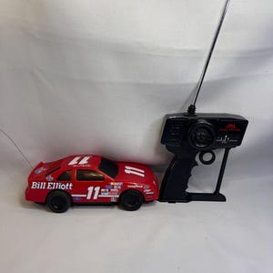 May include: A red radio-controlled race car featuring the number 11, the name Bill Elliott, and various sponsor decals, accompanied by a black handheld remote control unit with a long silver antenna and the text JRL Radio Control.