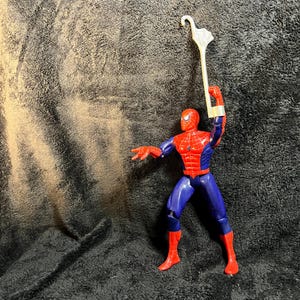 May include: A Spider-Man action figure in a dynamic pose, clad in a red and blue suit. The figure is holding a white, stylized umbrella. The figure's red mask and webbed design are clearly visible.