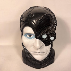 May include: A black and white plastic bust of a character with cybernetic enhancements. The figure has a stern expression, with one human eye and a cybernetic eye piece. The head is partially covered with black, mechanical components.