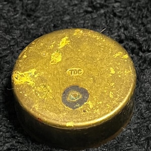 May include: A small, round, brass-colored object with a slightly tarnished appearance. The top surface has the letters "TDC" stamped into it. The object has a dark spot in the center and some yellow discoloration.