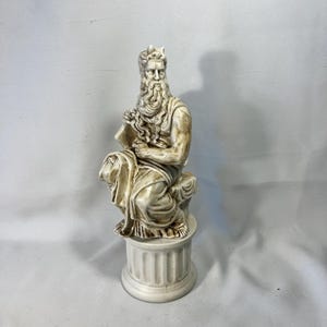 May include: A cream-colored statue of a seated figure with a long beard, holding a tablet. The figure is wearing a draped garment and a crown. The statue is mounted on a round, fluted pedestal.