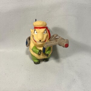 May include: A cartoonish sheep figurine with a stern expression, wearing a red headband and holding a tan weapon with a red tip. The sheep has a yellow body, green pants, and blue accents.