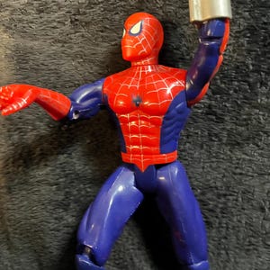 May include: A red, blue, and silver Spider-Man action figure. The figure has a red head and torso with a black spider emblem, blue legs and arms, and red hands and feet. The figure is holding a silver web shooter.