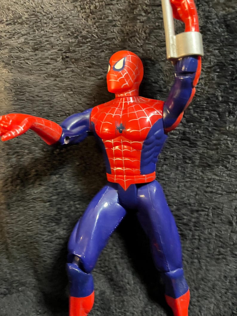 May include: A red, blue, and silver Spider-Man action figure. The figure has a red head and torso with a black spider emblem, blue legs and arms, and red hands and feet. The figure is holding a silver web shooter.