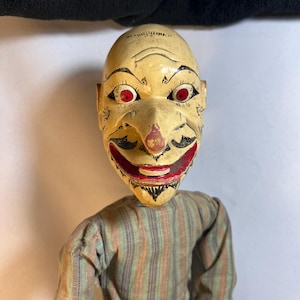 May include: A puppet with a carved, cream-colored wooden head featuring a pointed nose, red eyes, and a wide, painted red mouth with black facial details. The puppet wears a long-sleeved shirt made of fabric with vertical stripes in muted green, brown, and tan.