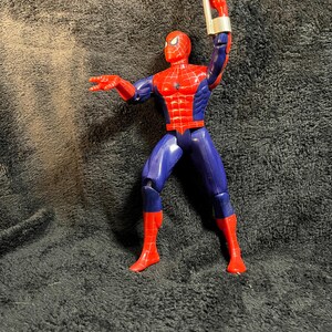 May include: A Spider-Man action figure in a dynamic pose, featuring a red and blue costume. The figure has a red mask and web detailing on the chest. It is holding a silver web-shooter accessory.