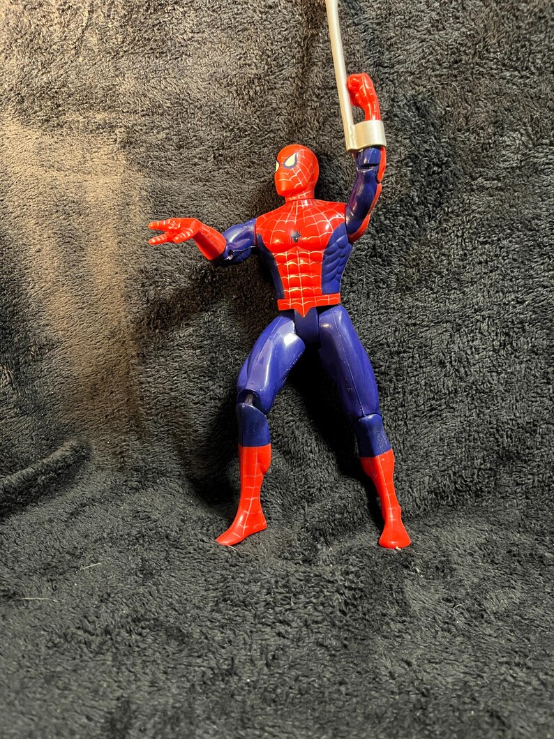 May include: A Spider-Man action figure in a dynamic pose, featuring a red and blue costume. The figure has a red mask and web detailing on the chest. It is holding a silver web-shooter accessory.