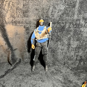 May include: Action figure of a character in a blue turtleneck, black pants, and boots. The figure wears a tactical vest and a mask with yellow lenses. It holds a weapon and has a cable attached.