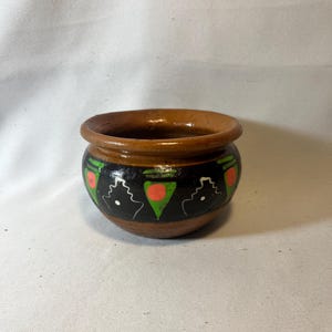May include: A small, round, brown ceramic pot with a black band featuring white, green, and red geometric designs. The rim of the pot is a darker brown, and the interior is glazed. The pot is likely handmade and suitable for small plants or decorative use.