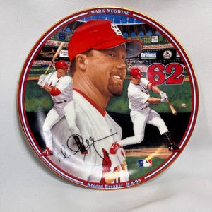 May include: A round commemorative collector plate featuring a portrait of baseball player Mark McGwire in a red cap, flanked by two images of him batting in a Cardinals uniform. The plate displays the number 62, a signature, and the text Record Breaker 9-8-98.
