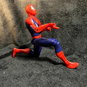 May include: A red and blue Spider-Man action figure in a crouching pose, pointing with one hand. The figure has a red head and feet, with a blue body and arms. The figure is on a dark surface.